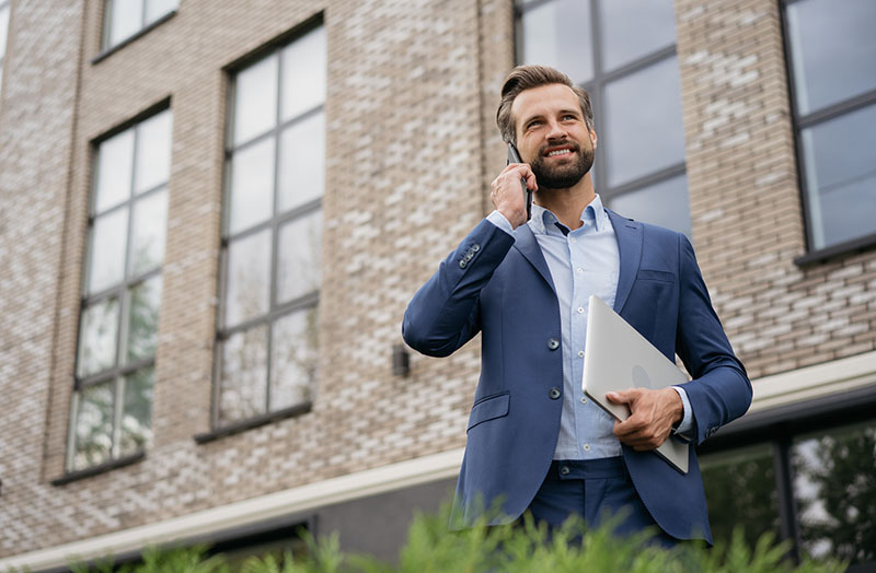 Handsome smiling businessman wearing stylish suit talking on mobile phone, holding laptop standing on the street. 